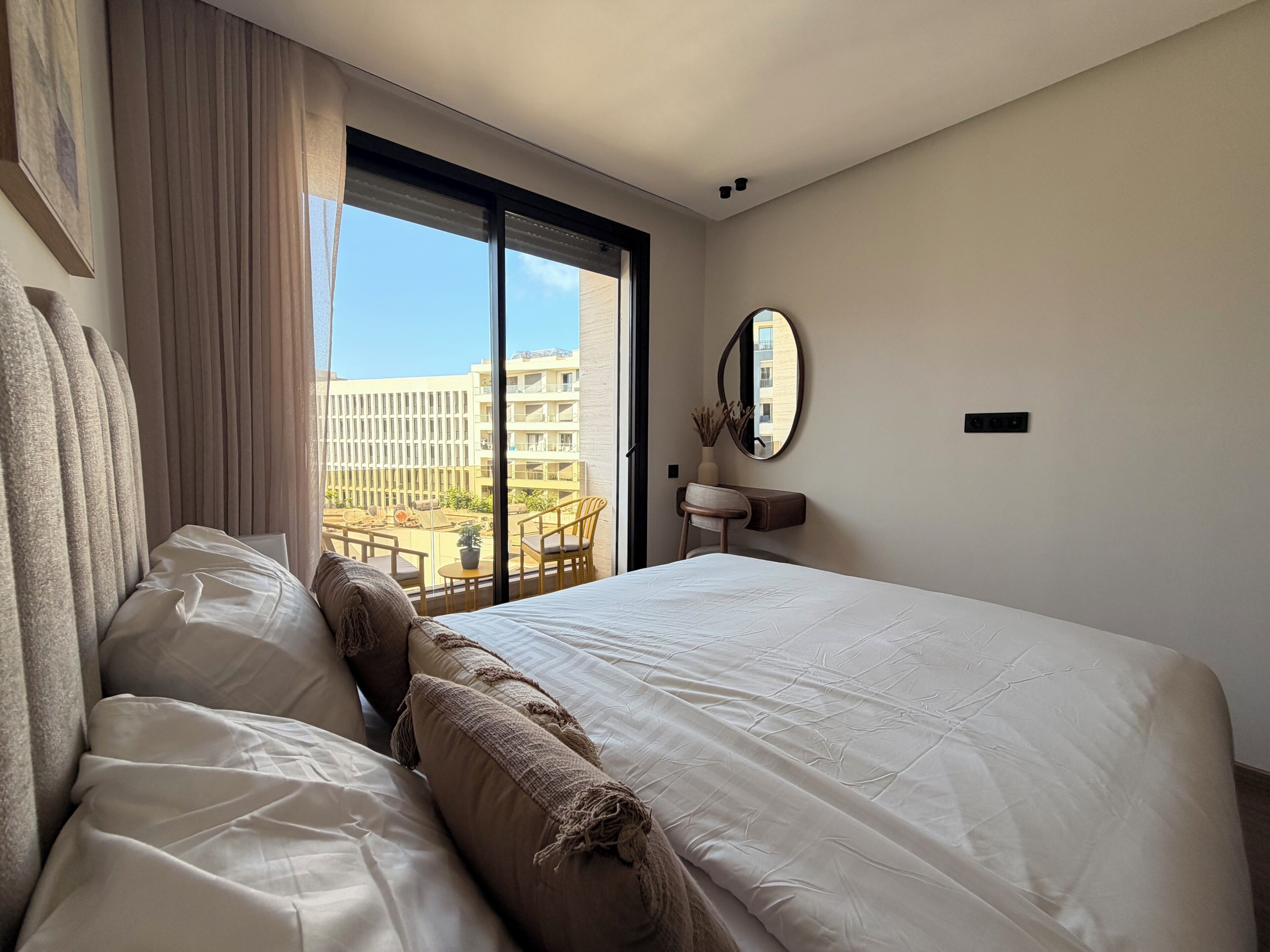 Habitación de hotel con una cama grande, un escritorio con silla, una ventana con cortinas y vistas a edificios.