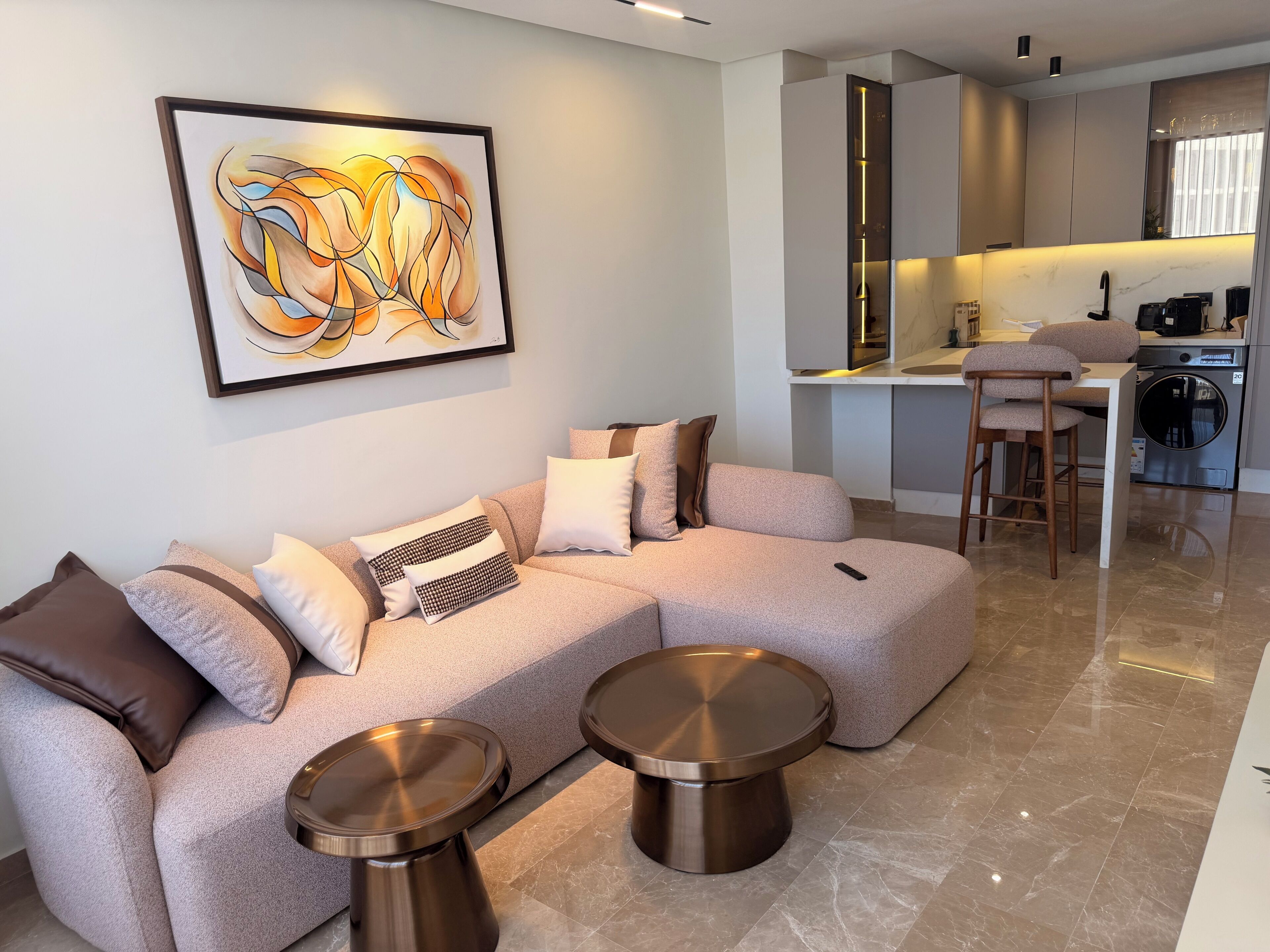 Una sala moderna con un sofá seccional, dos mesitas metálicas, una zona de cocina con un taburete alto y un cuadro abstracto grande en la pared.