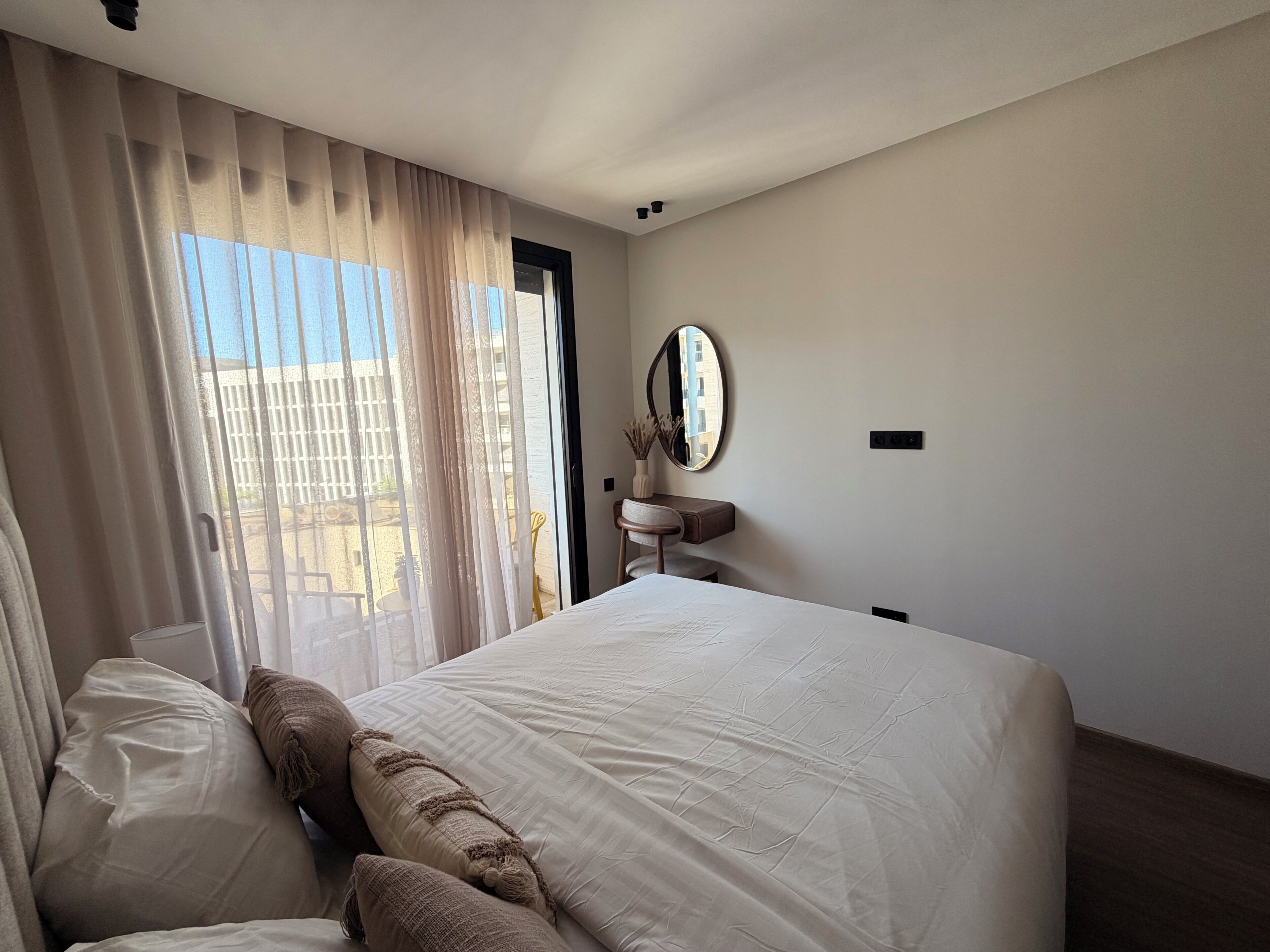 A hotel room with a large bed, a bedside table with a mirror, and a view of the outside.