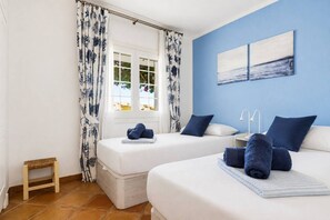 Ein Schlafzimmer mit zwei Betten, einem Fenster mit Vorhängen, einem Nachttisch mit Lampe und einem Kunstwerk an der Wand.