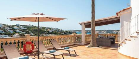 A rooftop terrace with a pool, lounge chairs, a sun umbrella, and a view of the coastline.
