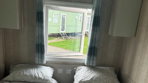 A room with two beds, a window with curtains, and a view of a green house outside. - Caravan Sleeps 8 With Sea Views and Parking (Morecambe)