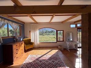 A room with a wooden ceiling, a large window with a view of a field, a yellow armchair, a wooden dresser, and a white desk with a mirror.