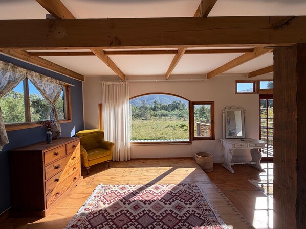 A room with a wooden ceiling, a large window with a view of a field, a yellow armchair, a wooden dresser, and a white desk with a mirror.