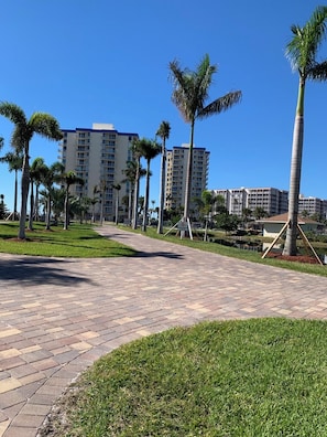 A paved walkway with palm trees, a grassy area, and high-rise buildings in the background. - NEW LISTING - 1 bedroom condo in FMB on the 7th floor with direct gulf views. (Fort Myers Beach)