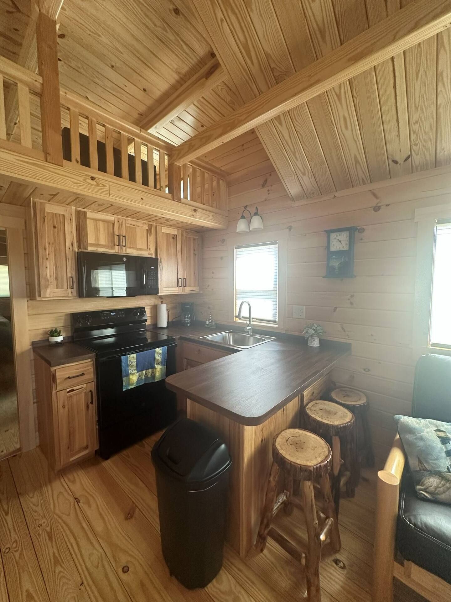A cabin-style kitchen with wooden cabinetry, a microwave, stove, and a sink. There are stools at the island and a trash can.