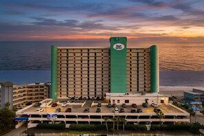 A hotel with a large parking lot, a prominent green vertical structure, and a view of the ocean at sunset. - Oceanfront Condo with Resort Pool, Hot Tub and Indoor Pool — Garden City Beach, (Garden City Beach)