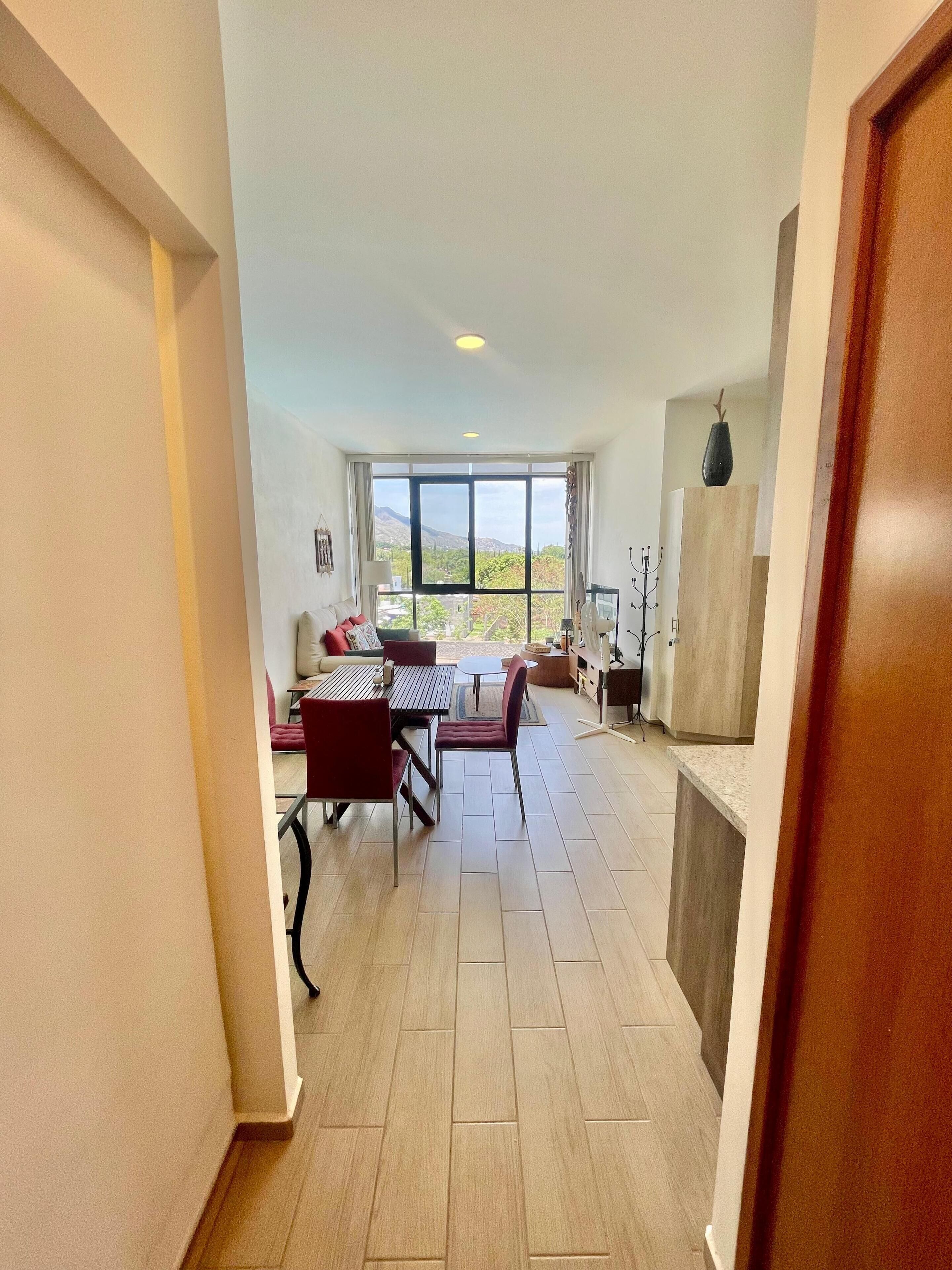 A spacious living area with a dining table and chairs, a sofa, and a view through large windows.