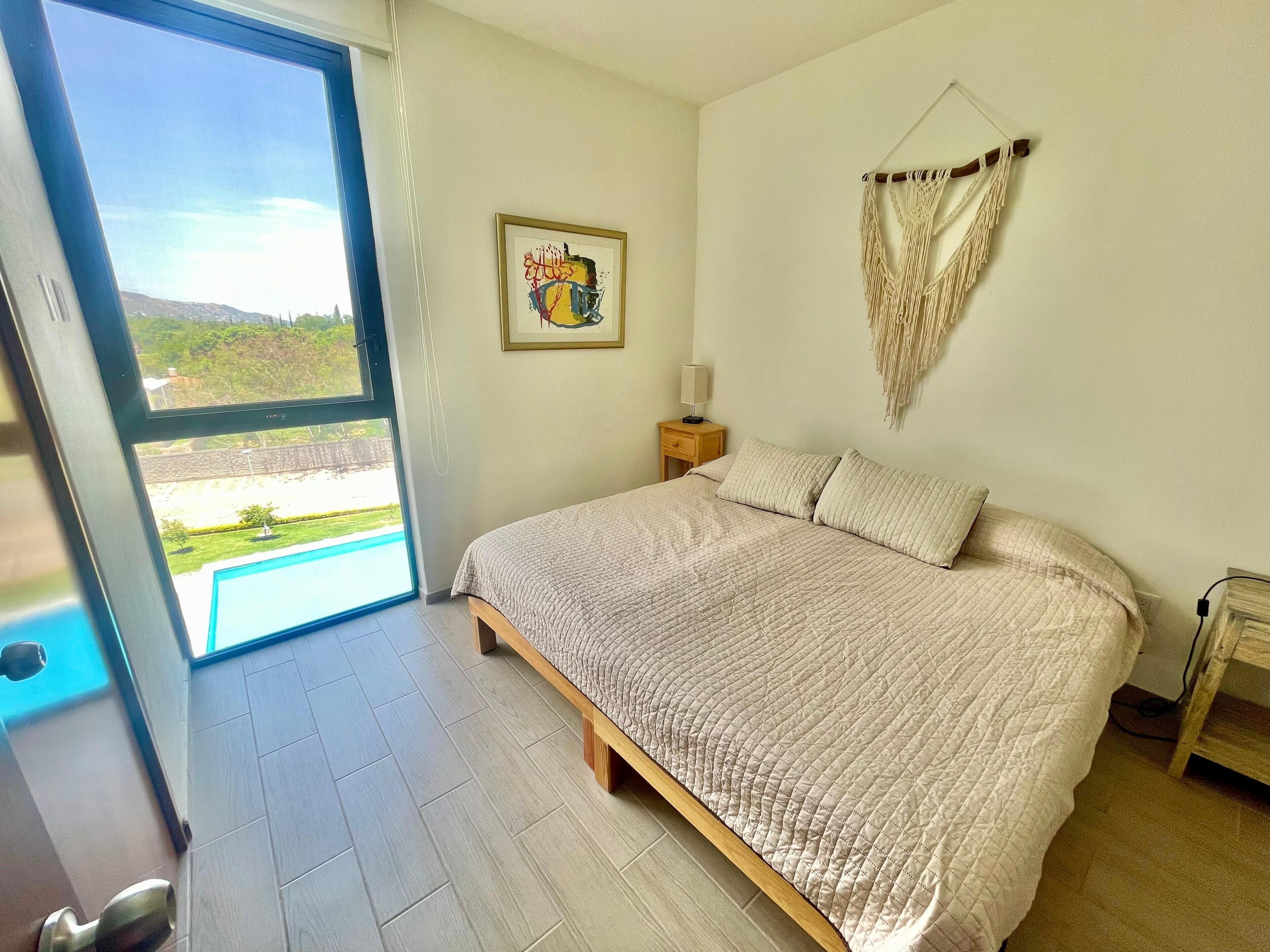 Un dormitorio con una cama grande, un ventanal con vista a una piscina, un tapiz de macramé y una obra de arte enmarcada.