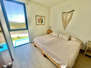 A bedroom with a large bed, a window with a view of a pool, a macrame wall hanging, and a framed artwork. - Apartment in El Chante Lake View (Chantepec)