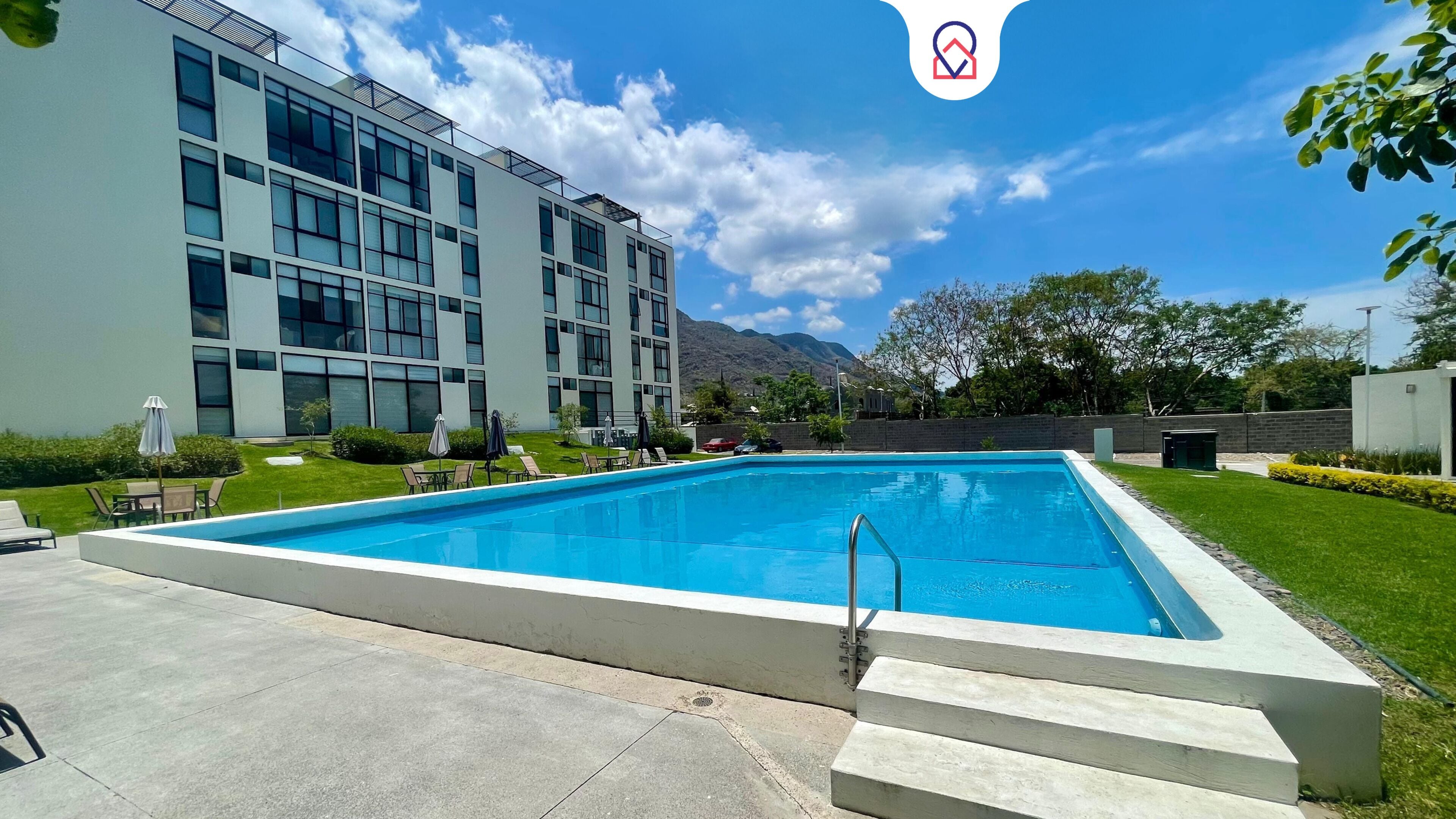 Una piscina rectangular con una escalera metálica, rodeada por un solárium de hormigón. Se observan sillones de descanso y sombrillas al fondo, y un edificio moderno con amplios ventanales.