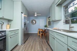 A kitchen with light green cabinets, a stainless steel refrigerator, a wooden dining table with chairs, a ceiling fan, and a window with a view of greenery. - Orange Grove home - fully renovated minutes from downtown (St. Augustine)