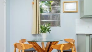 Uma área de refeições com mesa de vidro e cadeiras de madeira, uma planta em vaso, uma janela com cortinas e uma cozinha com armários.