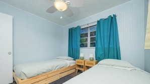 Quarto com duas camas de solteiro, ventoinha no teto, janela com cortinas e mesas de cabeceira com luminárias.