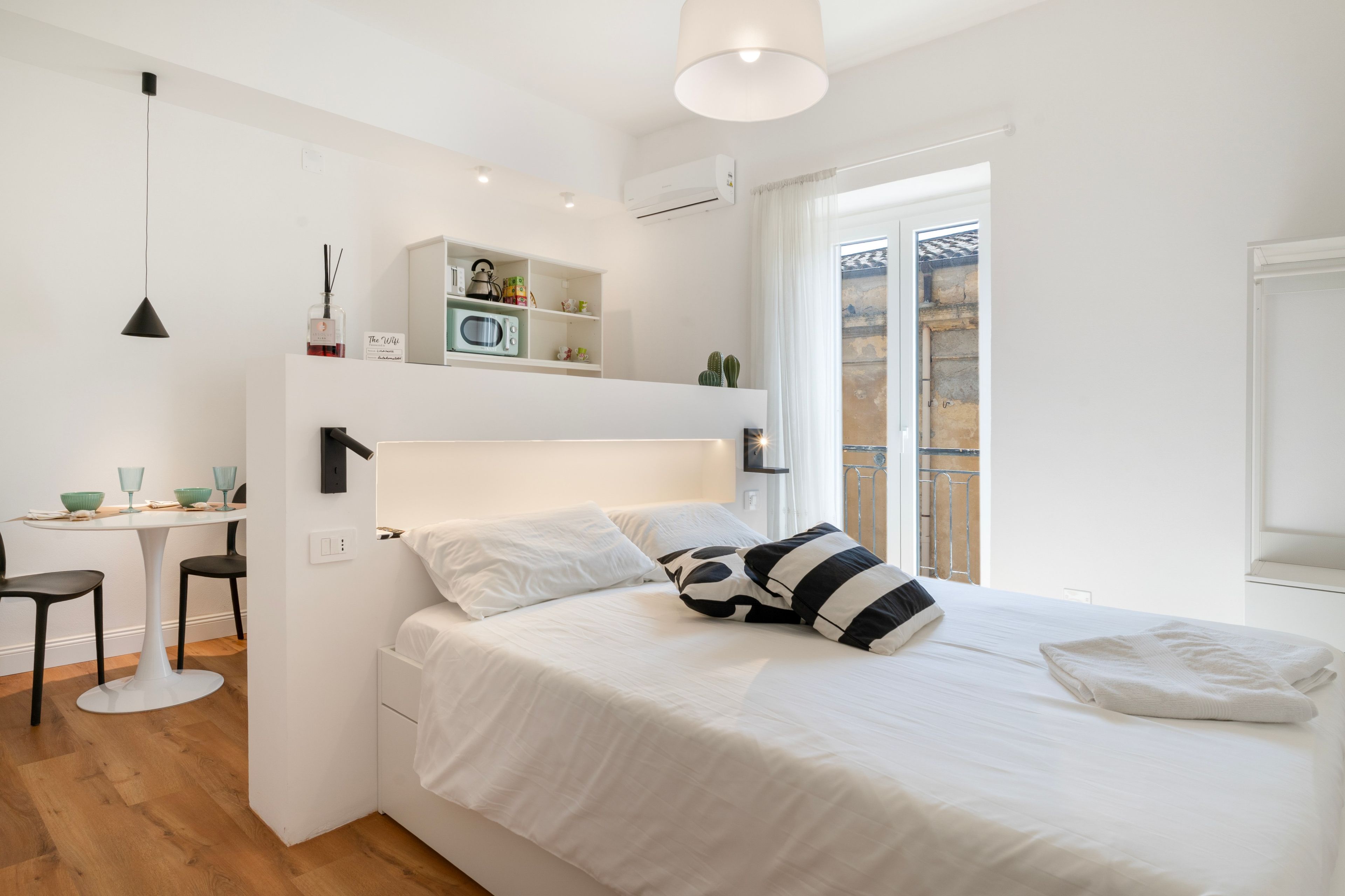 A modern bedroom with a bed, bedside table, and a small kitchenette area.