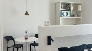 Ein modernes Zimmer mit einem weißen Bett, einem kleinen runden Tisch mit Stühlen, einer weißen Ablagefläche mit Küchengeräten und einer Pendelleuchte.