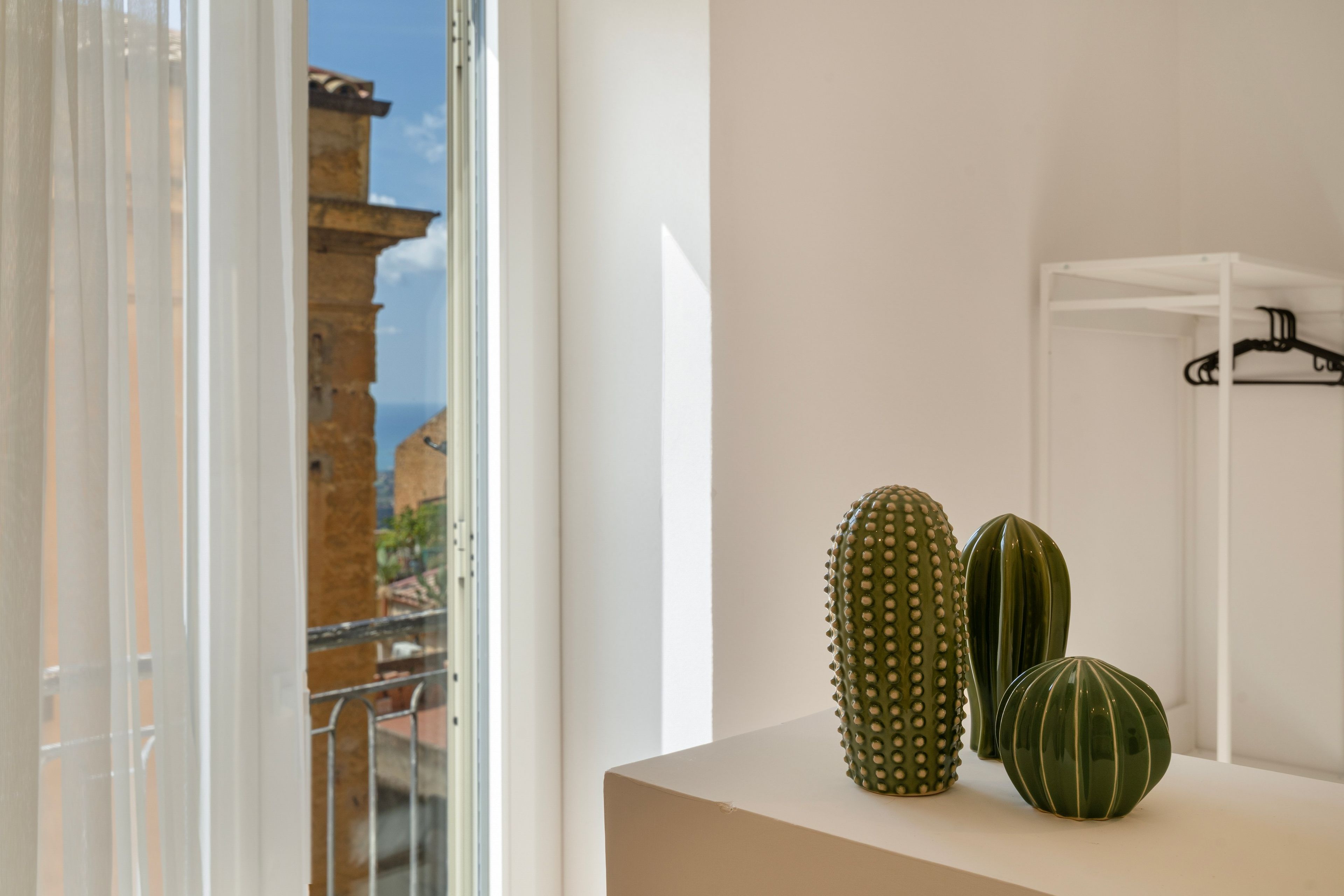 A room with a view of a building through a window, a white shelf with two green vases, and a white shelf with a black hanger.