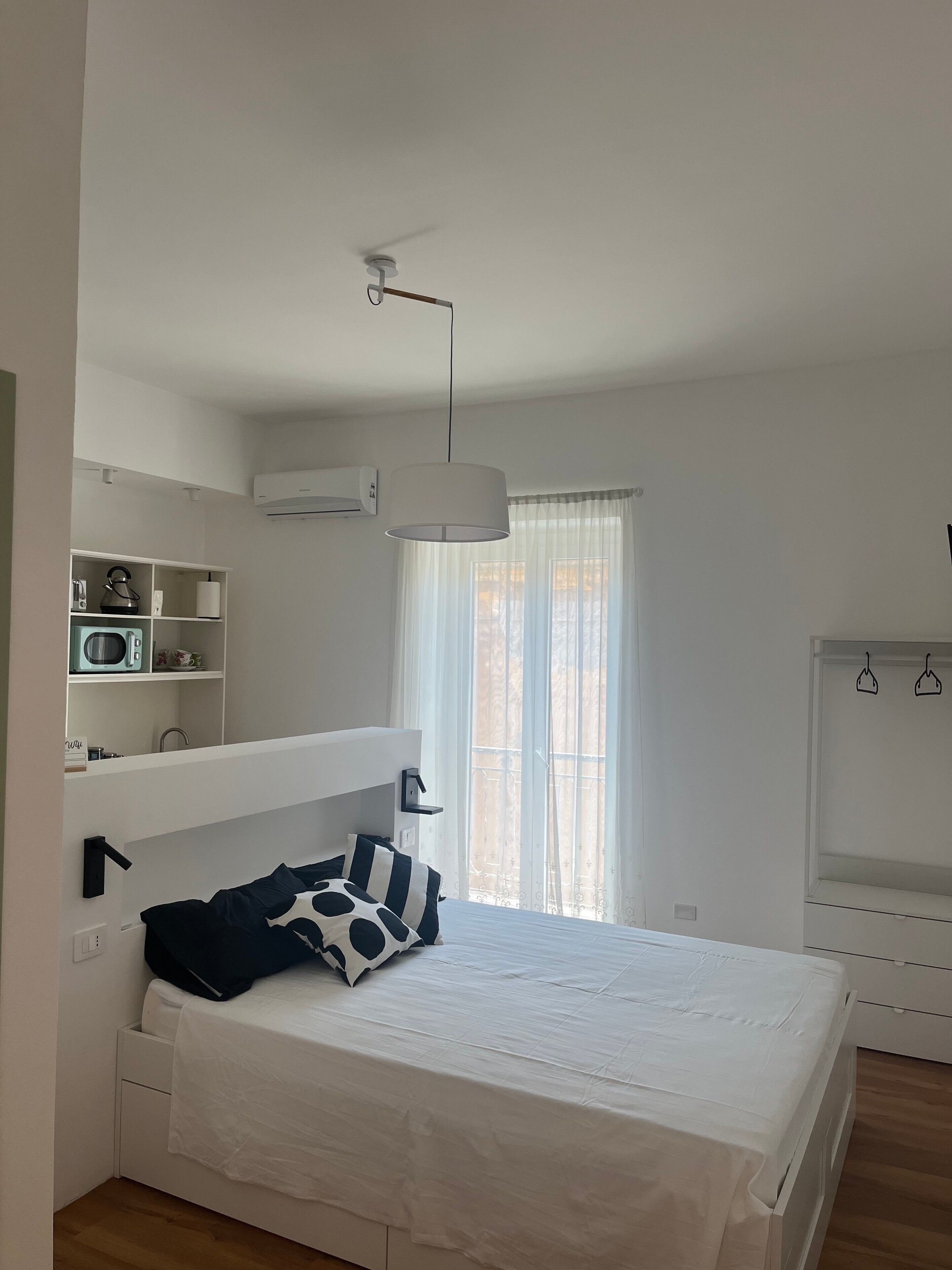 Una camera da letto con un letto, pareti bianche, una ventola a soffitto, una finestra con tende e un armadio bianco.