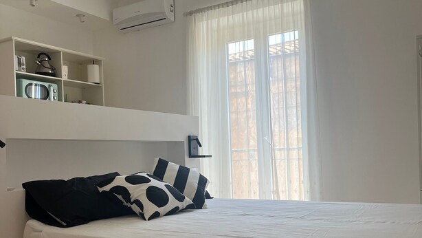 Ein Schlafzimmer mit Bett, einer Hängelampe, einer eingebauten Schrankwand mit Küchengeräten und einem Fenster mit durchsichtigen Vorhängen.