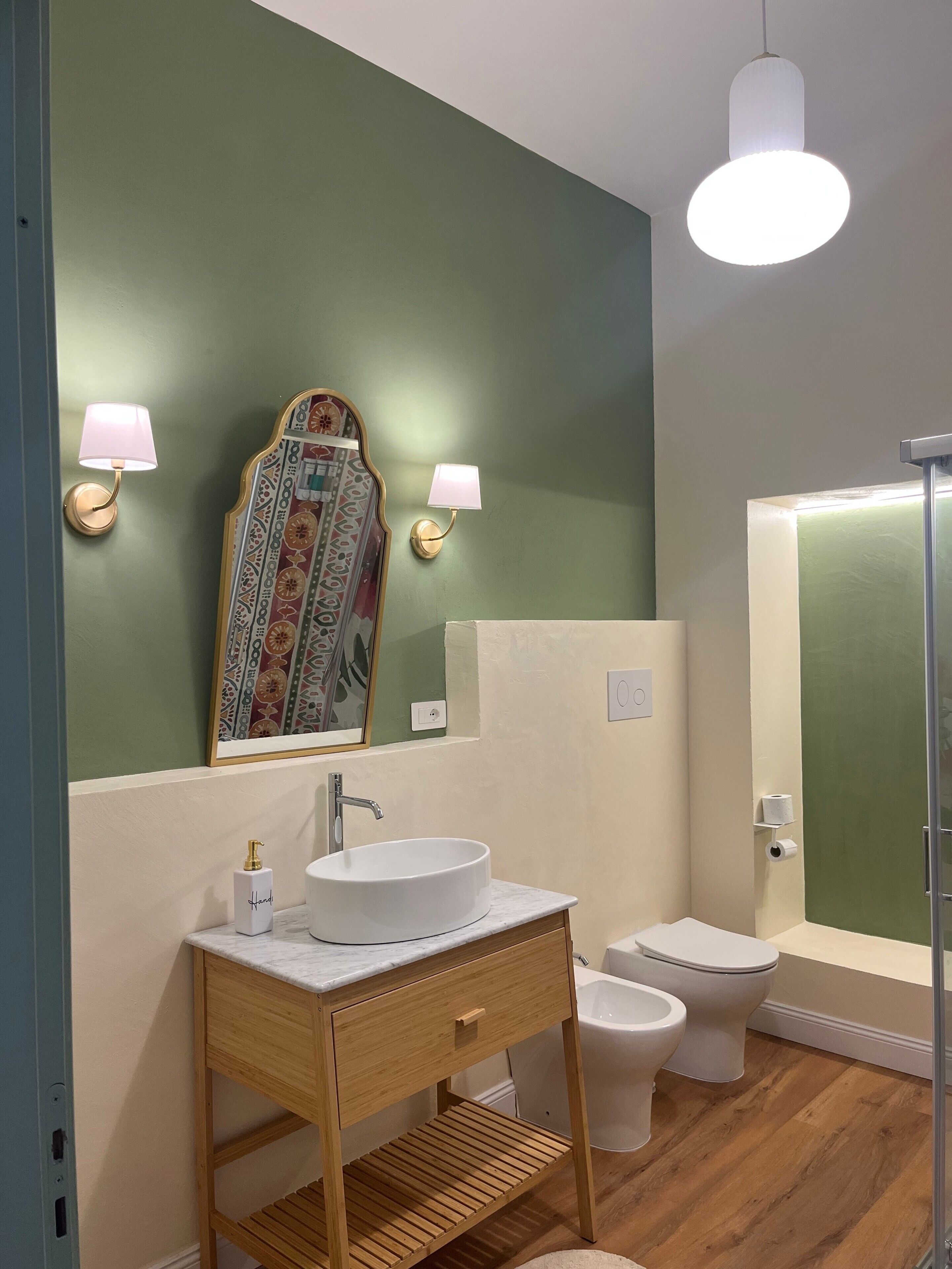 A bathroom with a white sink, two wall-mounted lights, a mirror with a decorative frame, a toilet, and a shower area.