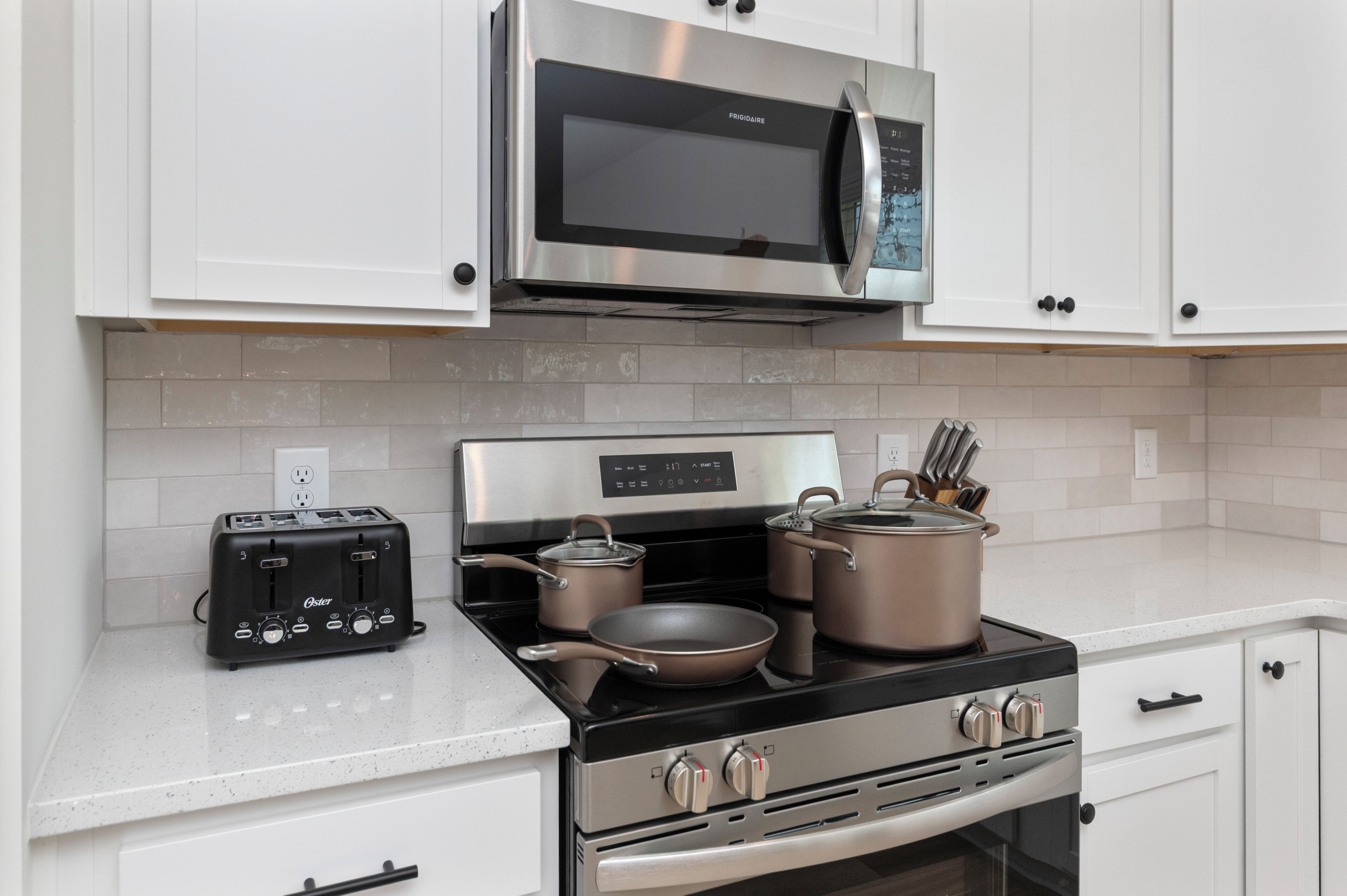 A modern kitchen with stainless steel appliances, including a microwave, toaster, and stove, with white cabinetry and a tiled backsplash.
