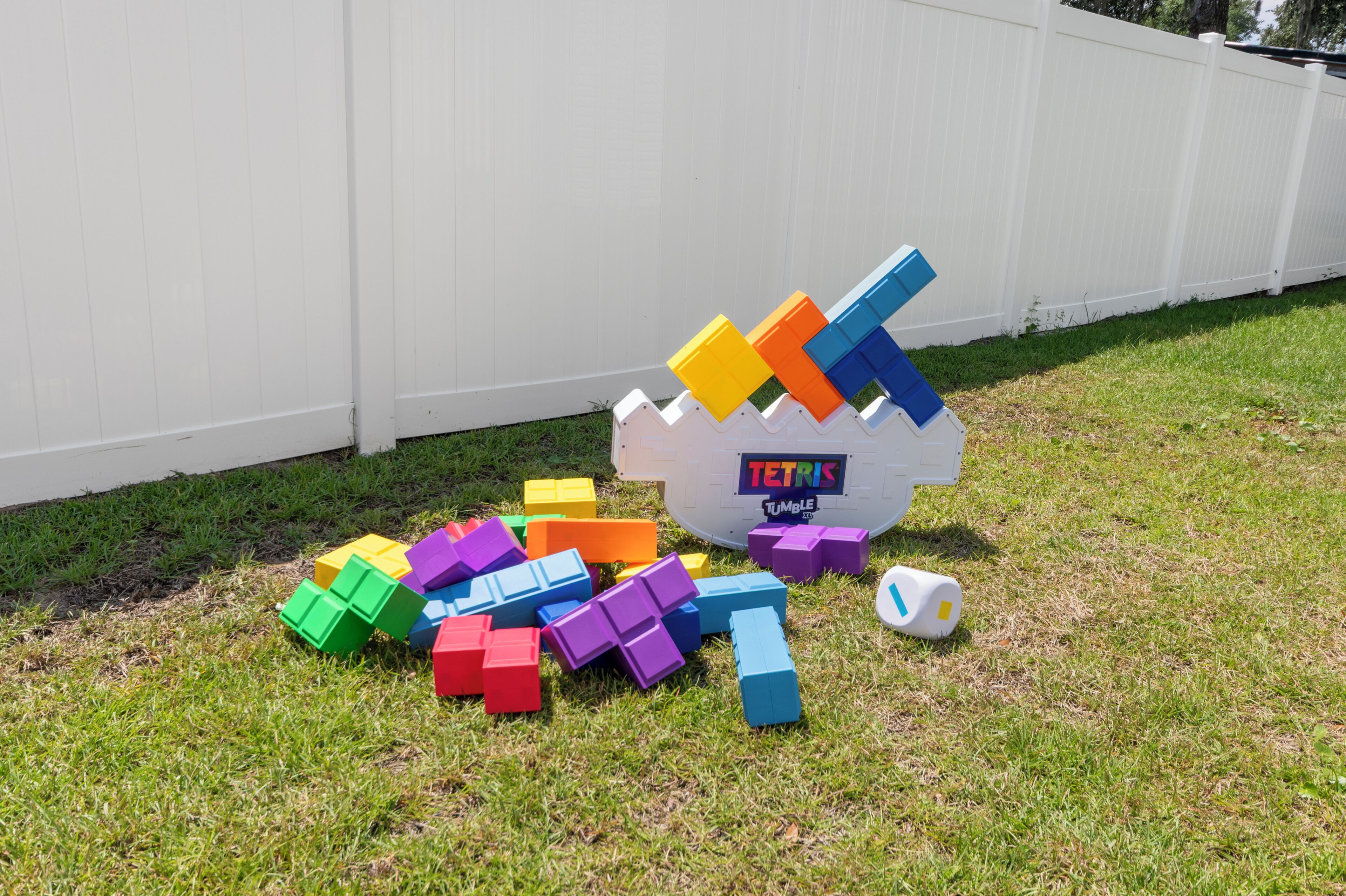 Ein weißer Zaun mit einem Tetris-Schild und bunten Blöcken, die auf dem Gras verteilt liegen.