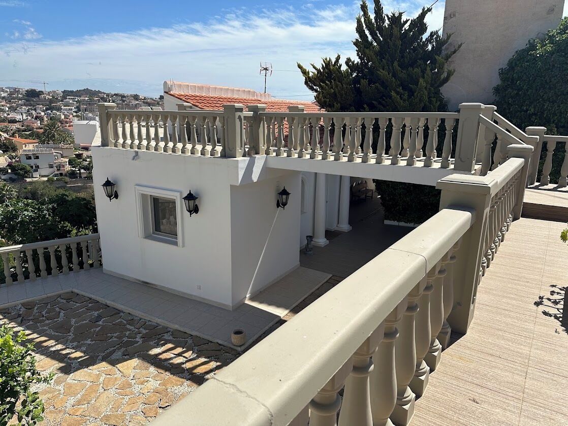 A balcony with a white railing, a small white building with a window, and a view of a town with red-roofed buildings.