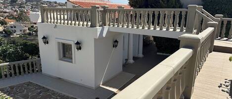 A balcony with a white railing, a small white building with a window, and a view of a town with red-roofed buildings.