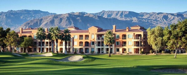 Een meerhoog hotel met een golfbaan ervoor, omgeven door bergen.
