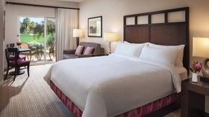 Hotelkamer met een groot bed, een bureau met stoel, een bank en uitzicht op een golfbaan door een schuifdeur.