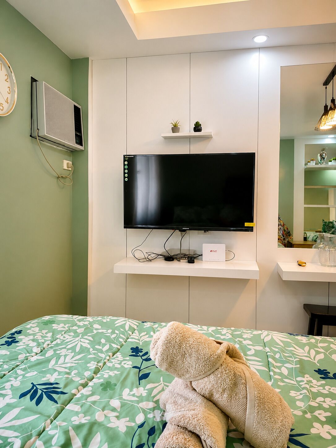 Ein Schlafzimmer mit einem Bett, einem an der Wand montierten Fernseher, einer kleinen Ablage mit Pflanzen und einem Spiegel über dem Bett.
