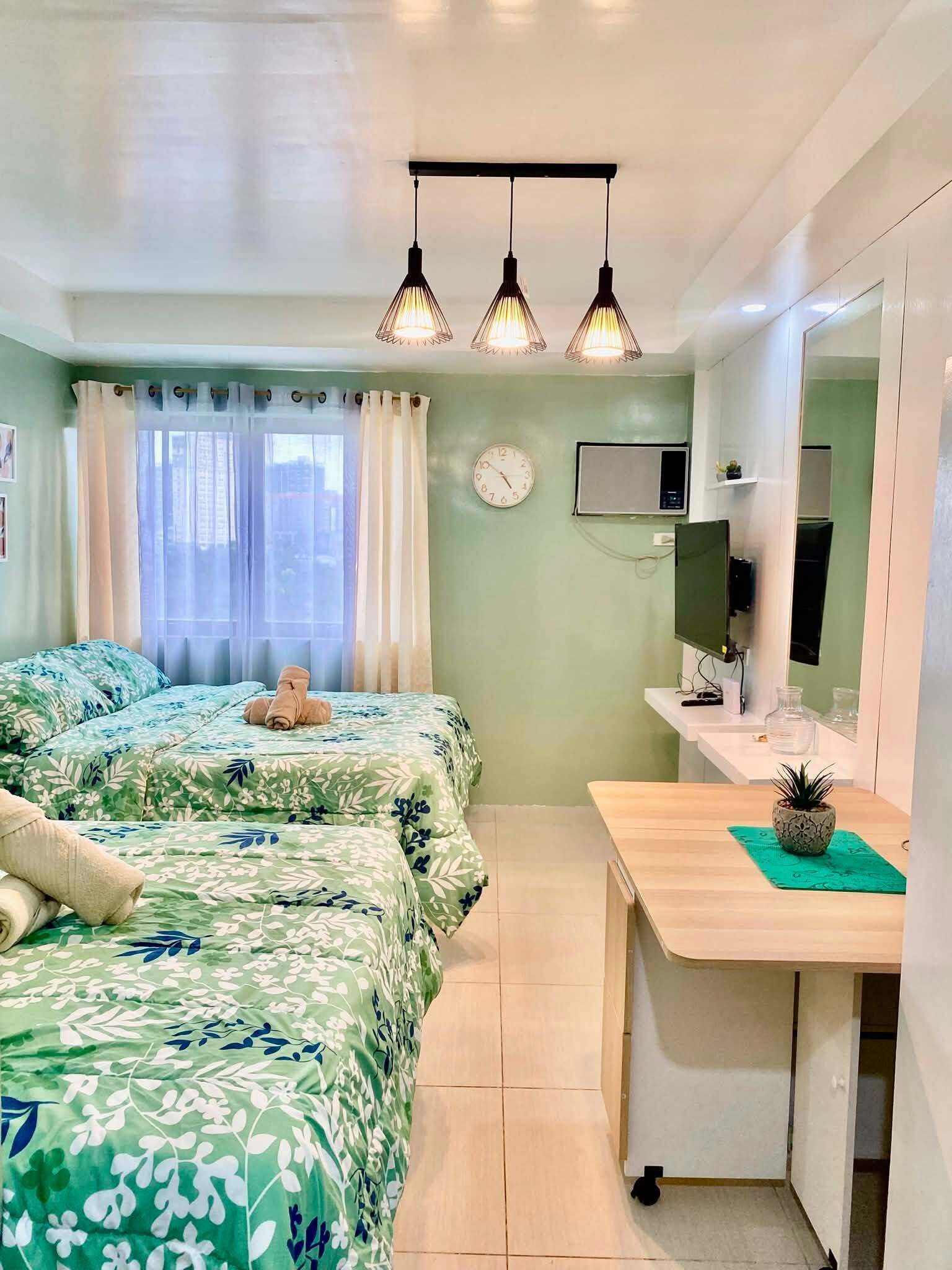 Quarto de hotel com duas camas, uma escrivaninha, um espelho, um relógio e uma janela com cortinas.