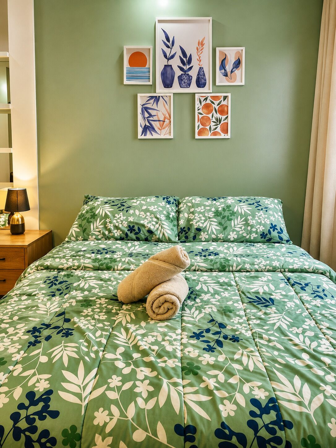 Een slaapkamer met een bedovertrek met groene bloemen, een opgerolde handdoek op het bed, en verschillende ingelijste kunstwerken aan de muur.