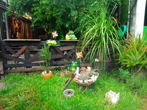 Un jardín con valla de madera, diferentes plantas en macetas, una estatua de rana y un pequeño carretilla.