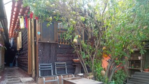 Uma área de estar ao ar livre rústica, com mesa e cadeiras de madeira, rodeada por um jardim com uma árvore e diversas plantas.