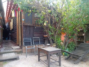 Een rustieke buitenruimte met een houten tafel en stoelen, omgeven door een tuin met een boom en diverse planten.