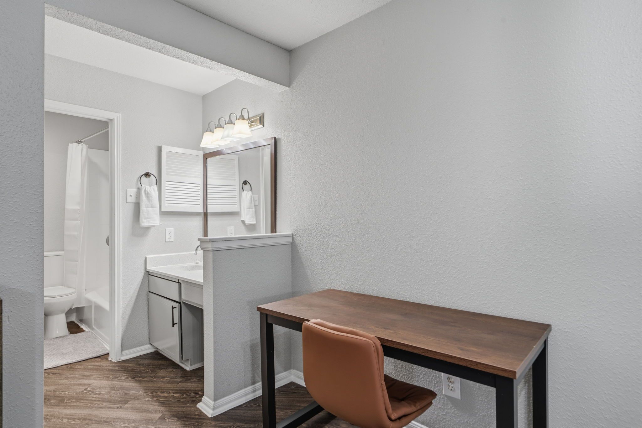 A compact bathroom with a shower, toilet, and sink. There's a small table with a chair in the corner.