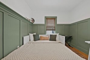 Een slaapkamer met een bed, kussens, een radiator, een raam met zonwering, en een klein tafeltje met een toetsenbord.