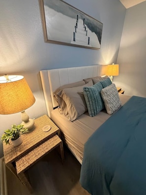Quarto de hotel com cama, duas luminárias de cabeceira, uma mesinha com uma planta e uma obra de arte na parede.