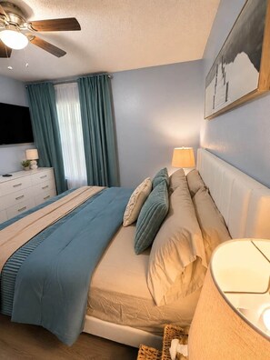 Una camera da letto con un letto, un comodino, una lampada, una ventola a soffitto, una TV e una finestra con tende.