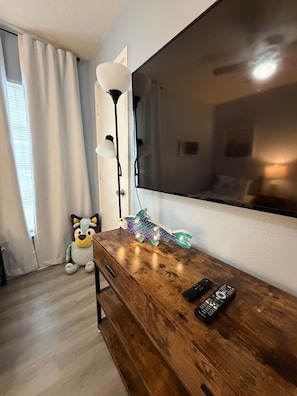 Quarto com cômoda de madeira, televisão de tela plana grande fixada na parede, um brinquedo de pelúcia e uma luminária decorativa em formato de peixe.