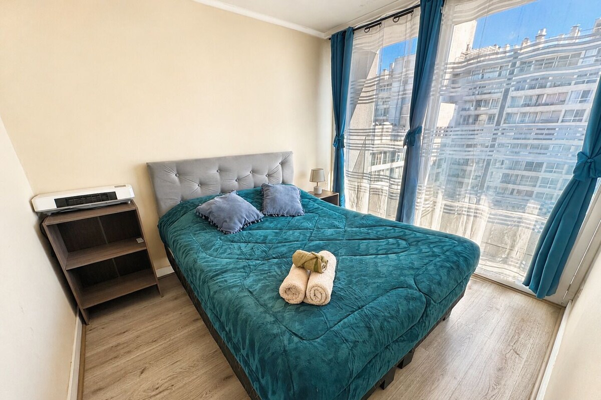 Une chambre à coucher avec un grand lit, une table de chevet, un radiateur et une vue sur le paysage urbain à travers des rideaux transparents.