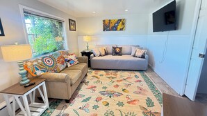 A living room with a beige sofa, colorful pillows, a patterned rug, a small side table, and a television mounted on the wall.