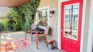 A wooden deck with two chairs and a floral rug, a red door, and a wall covered with green ivy.