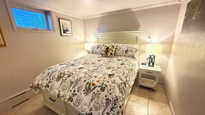 A bedroom with a floral bedspread, a nightstand with a lamp, a wall clock, and a window with blinds.