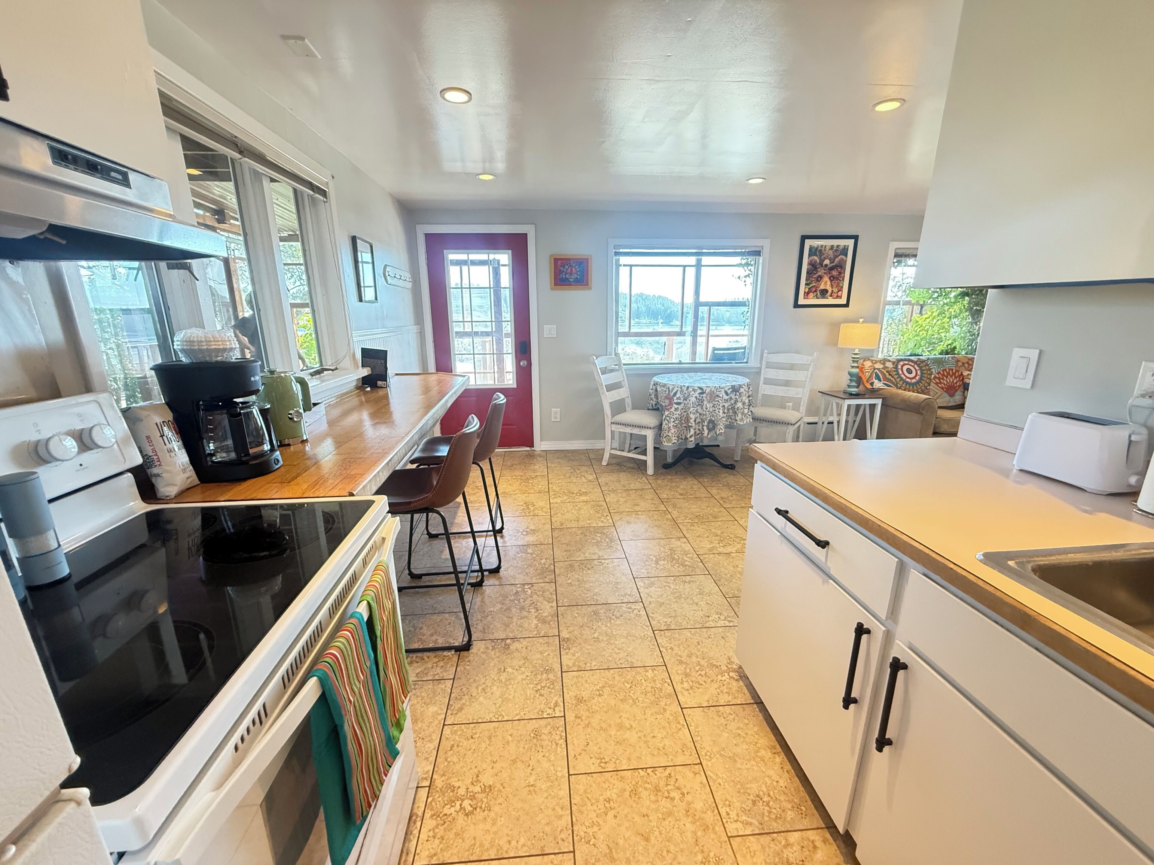 A kitchen with a stove, coffee maker, and chairs. There is a dining table with chairs and a window.