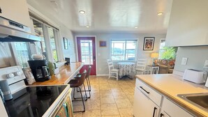 A kitchen with a stove, coffee maker, and chairs. There is a dining table with chairs and a window.