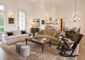 Un salon spacieux avec un canapé, une table basse, des poufs et un fauteuil à bascule. On aperçoit une cuisine au fond.