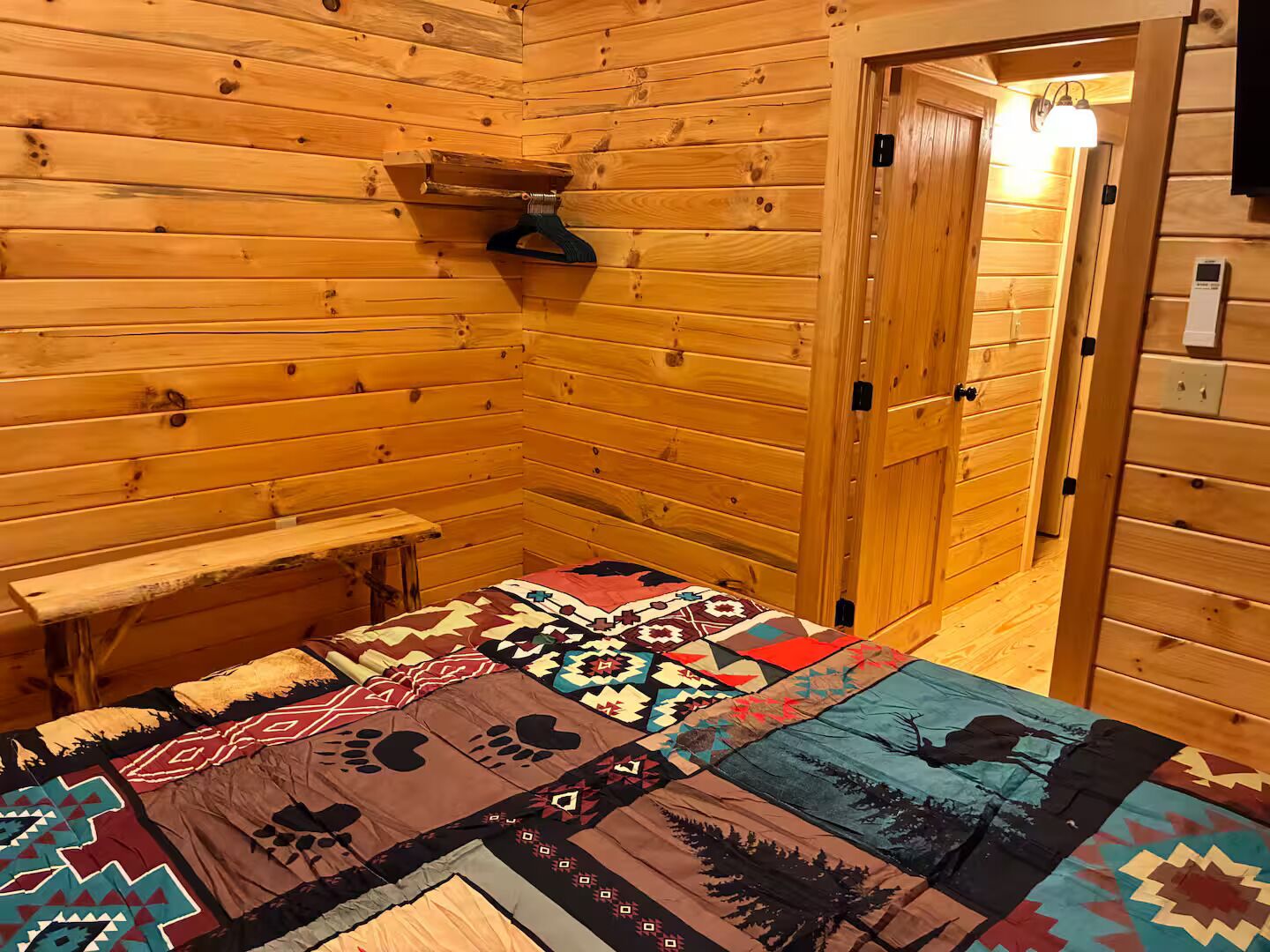 A wooden cabin interior with a bed, a bench, and a door leading outside.