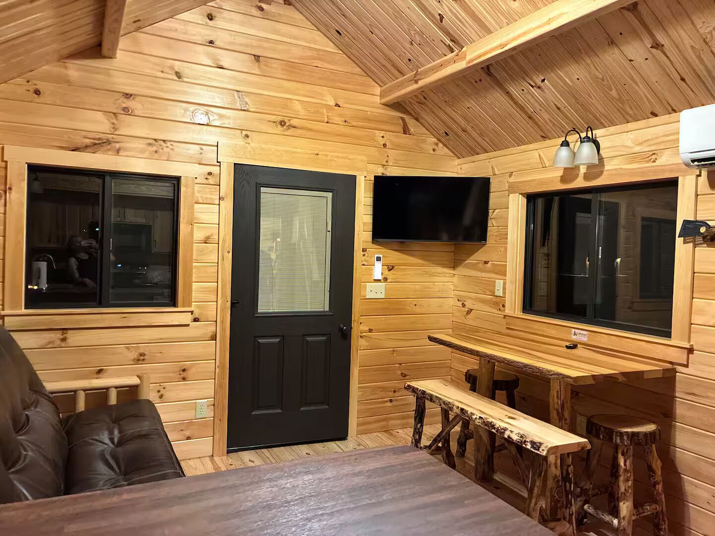 A wooden cabin interior with a black door, a television mounted on the wall, and a bench.